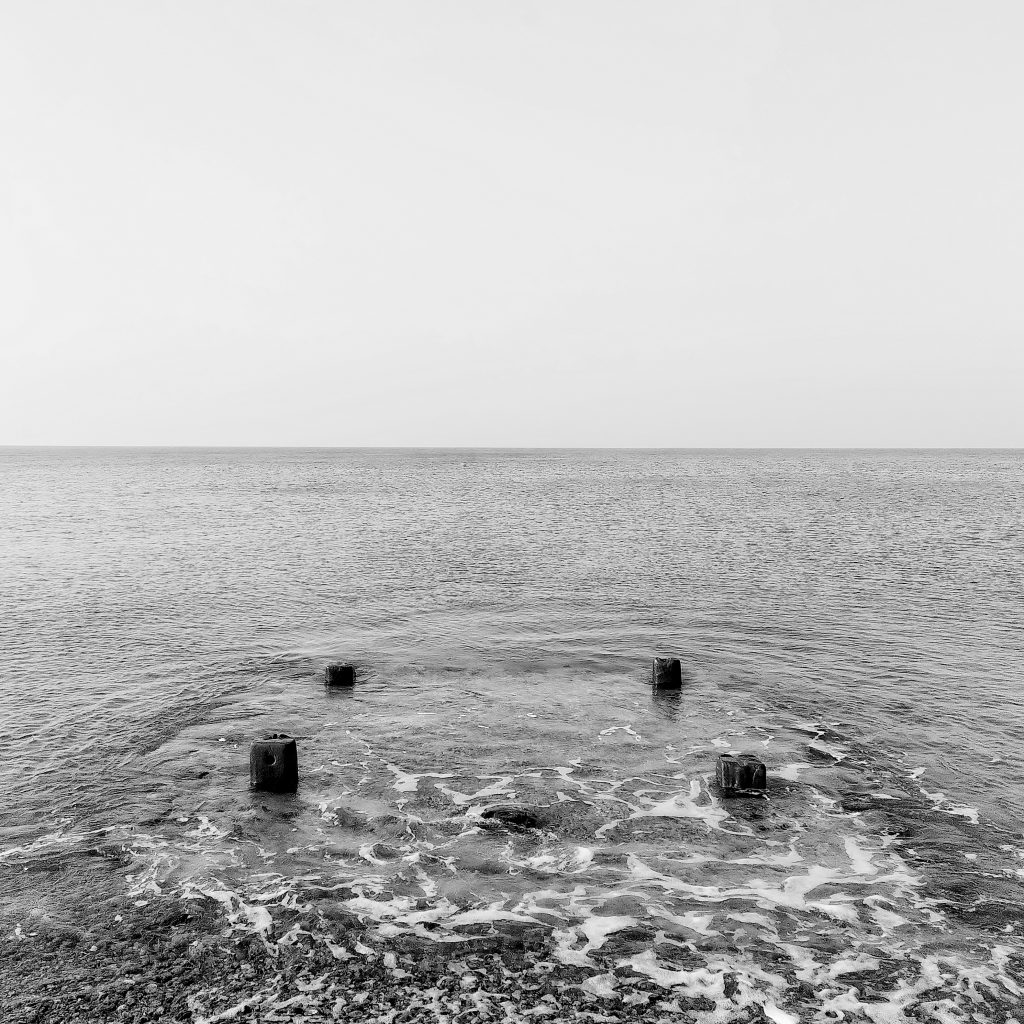 Black and white photo of the sea, with four old wooden pillars just protruding from the gentle waves.