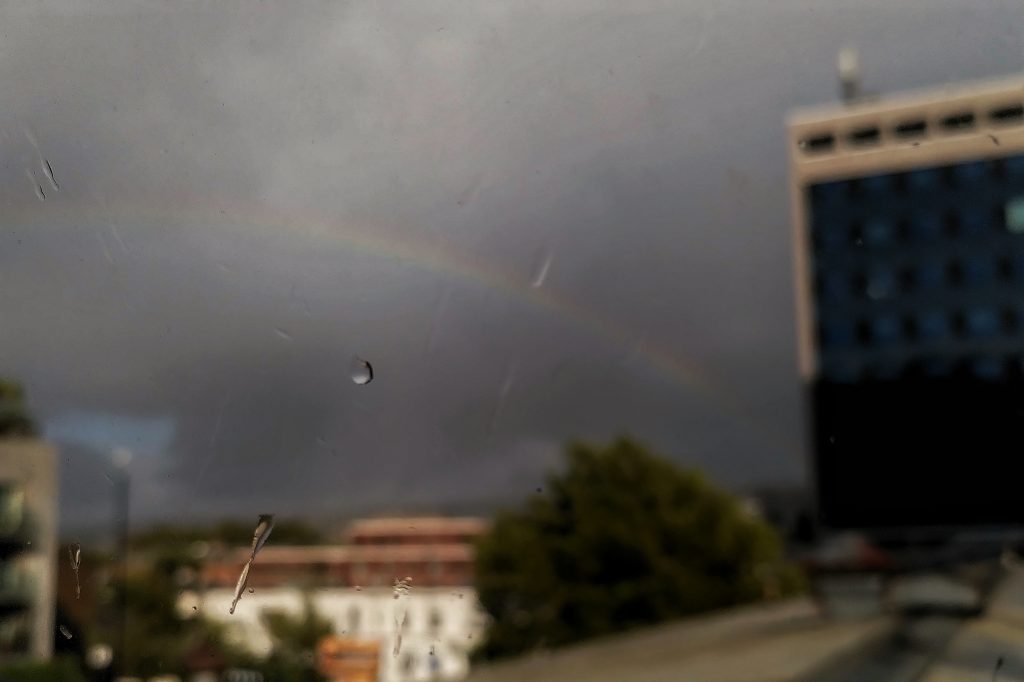 Photo of a raindrop on a window pane, with a blurred large building and a rainbow in the background.