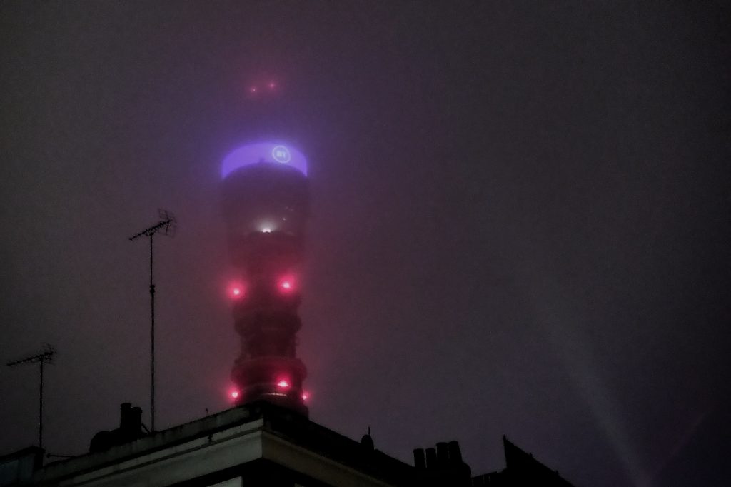 The BT tower in London in the dark, seen through a layer of fog. Sihouettes of a TV aerial and buildings are in the foreground.