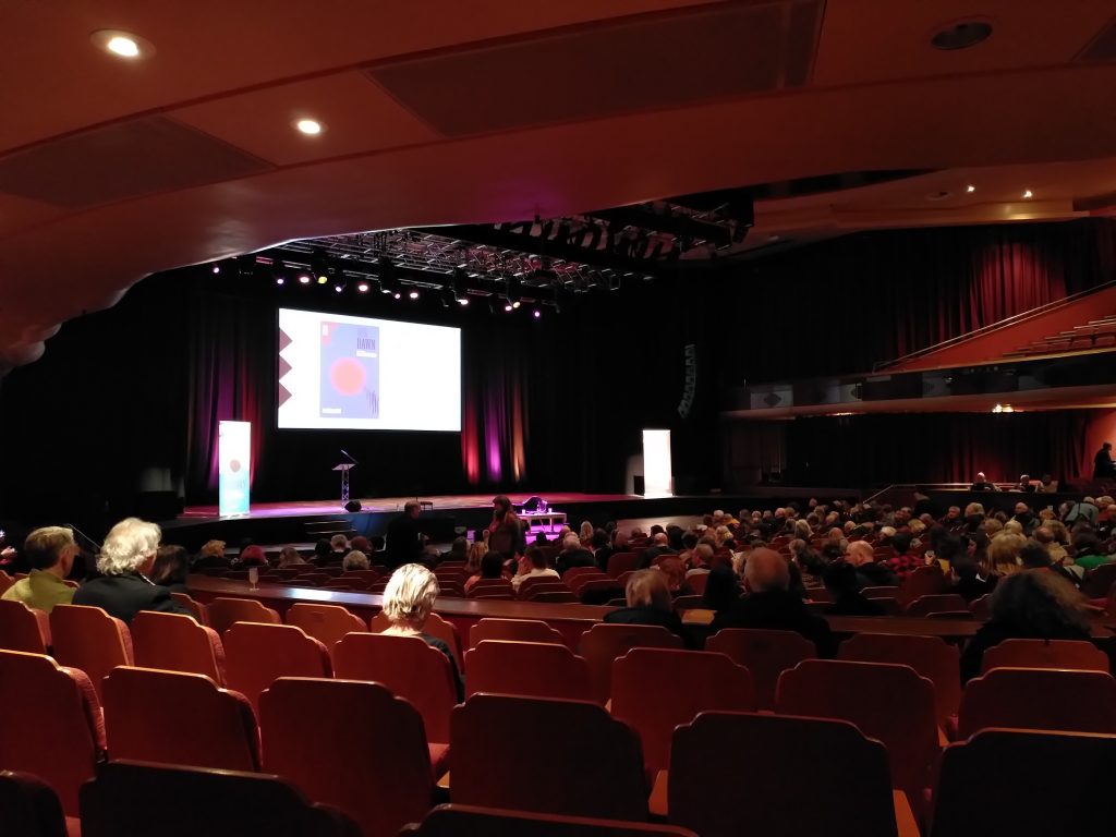 Inside of the Brighton Dome, a womb-like decor of dark red with seats stretching out in front, lots of heads, all facing towards a stage with a bright projection waiting for talks to start.