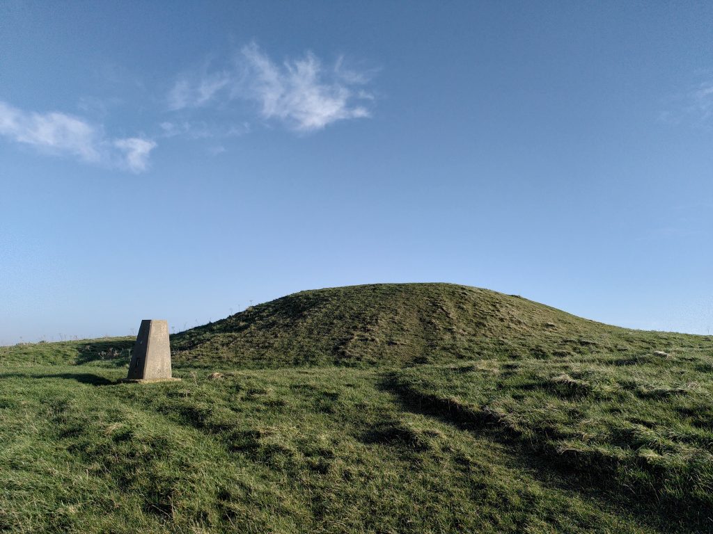 Photo looking up at a grassy burial mound with a stone pillar trigonometry point beside it, under a blue sky with a few clouds.