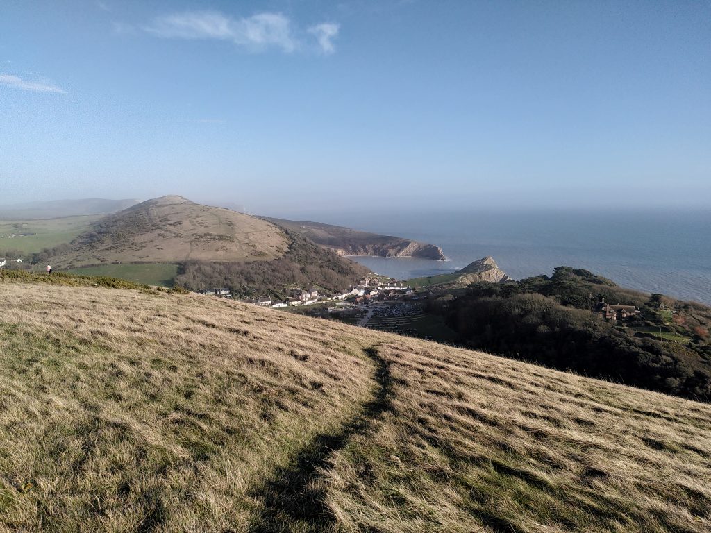 A view from the top of a hill, looking down to Lulworth Cove ithe distance below a blue sky.