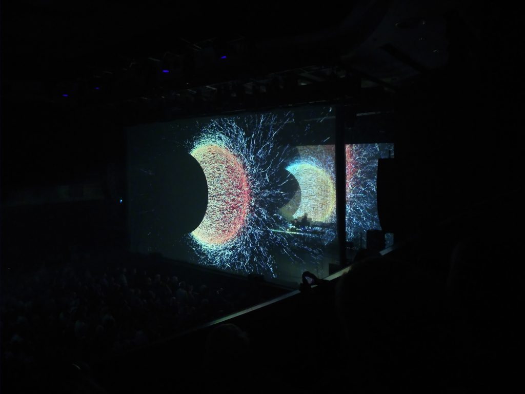 A stage n the dark. A giant sun-like projection is shown both in front of and behind the stage.