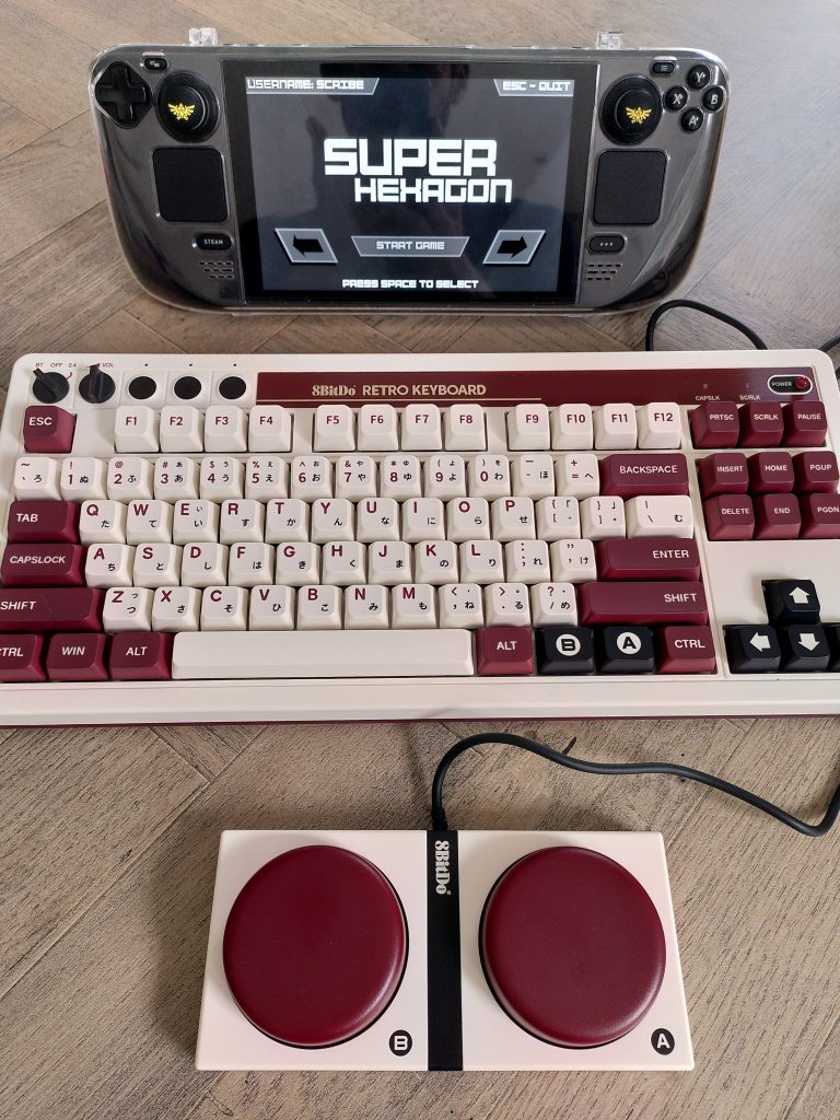 A cream and maroon-styled mechanical 8bitdo keyboard, with twolarge matching buttons in front, and a Steam Deck showing Super Hexagon in the background.