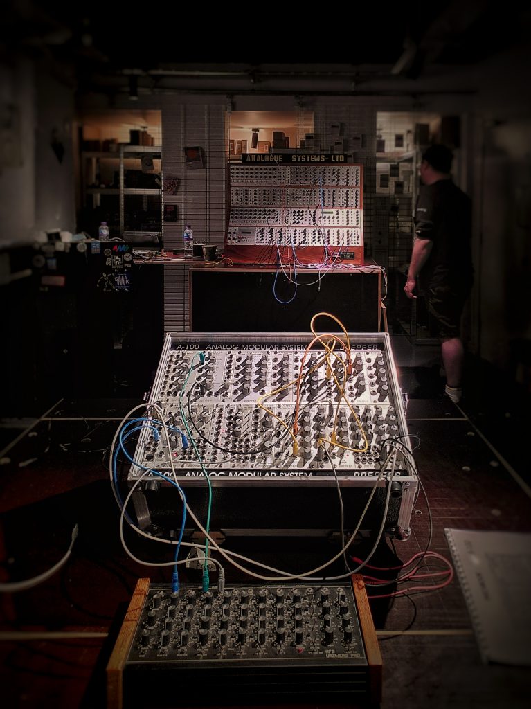 Several banks of modular synths, all dials and cables, sit in a dark room.