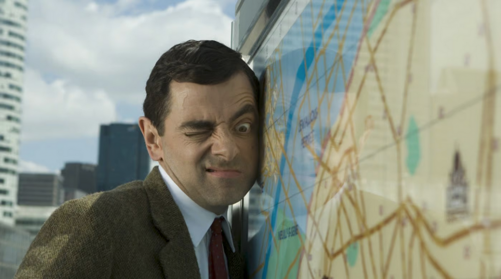 Mr Bean with his face pressed up against a map, one eye closed and the other staring down the map.
