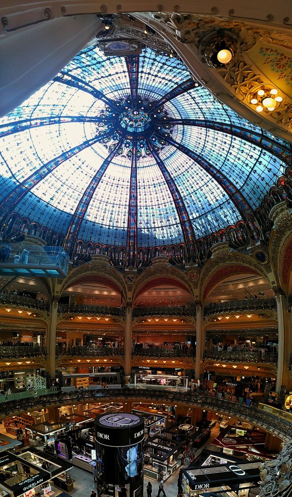 Photo showing the magnificent inner space of the Galeries Lafayette in Paris, with huge ornate arches and a giant glass dome towering over 3 floors of posh shops.