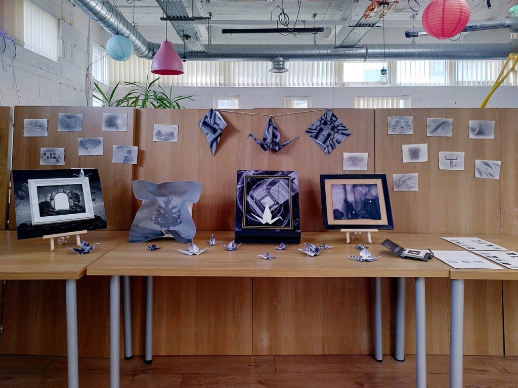Photo showing a range of framed and folded black and white art propped up against wood-effect desks inside a white room.